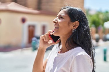 Genç, güzel, İspanyol bir kadın gülümsüyor ve caddede akıllı telefondan konuşuyor.