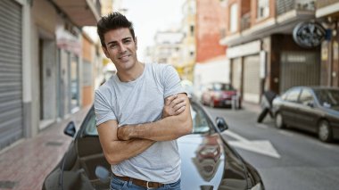 Kollarını kavuşturup caddeye gülümseyen Latin asıllı genç adam arabanın yanında duruyor.