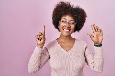 Genç Afro-Amerikalı kadın pembe arka planda duruyor ve altı numaralı parmaklarıyla işaret ederken kendinden emin ve mutlu gülümsüyor. 