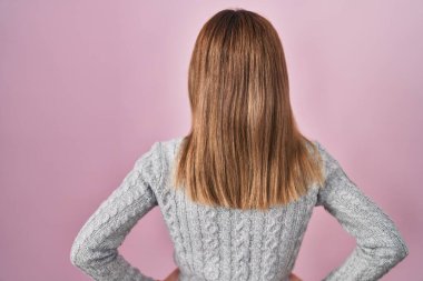 Güzel bir kadın pembe arka planda dikiliyor geriye bakıyor kolları vücudunda. 