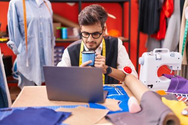 Genç İspanyol terzi atölyede kahve içerken dizüstü bilgisayar kullanıyor.