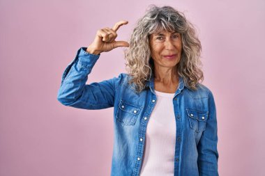 Middle age woman standing over pink background smiling and confident gesturing with hand doing small size sign with fingers looking and the camera. measure concept. 