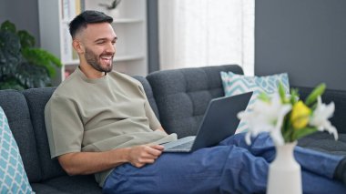 Genç İspanyol adam evdeki koltukta dizüstü bilgisayar kullanıyor.