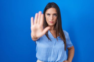 Young brunette woman standing over blue background doing stop sing with palm of the hand. warning expression with negative and serious gesture on the face. 