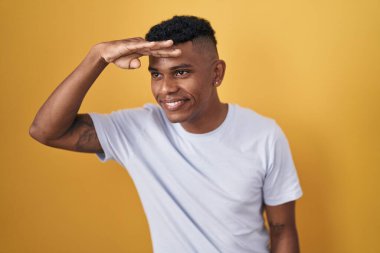 Young hispanic man standing over yellow background very happy and smiling looking far away with hand over head. searching concept. 