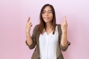 Orta yaşlı Çinli kadın pembe arka planda gözlük takıyor umut ve gözlerin kapalı bir şekilde gülümseyerek işaret ediyor. Şans ve batıl inançlar. 