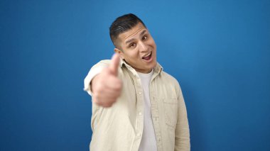 Genç İspanyol adam gülümsüyor. Kendine güveni tam. Başparmağıyla izole edilmiş mavi arkaplanı işaretle.