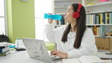 Laptop ve kulaklık kullanan genç, güzel İspanyol kız öğrenci üniversite sınıfında enerji içeceği içiyor.