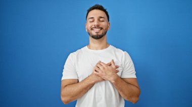 Genç Arap adam, elleri kalbimde, mavi arka planda, kendinden emin gülümsüyor.