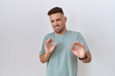 Hispanic man with beard standing over white background disgusted expression, displeased and fearful doing disgust face because aversion reaction. 