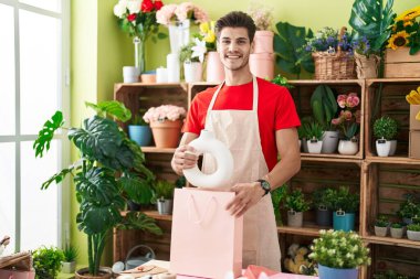 Young hispanic man florist smiling confident putting plant jar on shopping bag at flower shop