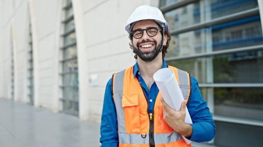 Genç İspanyol mimar. İnşaat alanının planlarını elinde tutarak gülümsüyor.