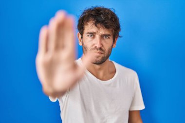 Hispanic young man standing over blue background doing stop sing with palm of the hand. warning expression with negative and serious gesture on the face. 