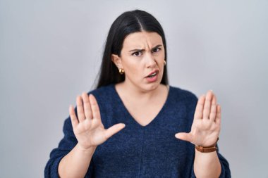 Young brunette woman standing over isolated background moving away hands palms showing refusal and denial with afraid and disgusting expression. stop and forbidden. 