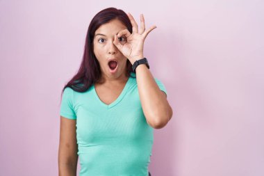 Pembe arka planda duran genç İspanyol kadın şaşırmış yüz ifadesiyle şaşırmış, parmaklarının arasından bakan bir jest yapıyor. İnançsız ifade. 