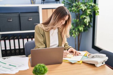 Young woman business worker using laptop writing on reminder paper at office