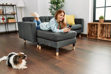 Beyaz kadın akıllı telefon kullanıyor. Evdeki kanepede yatıyor.