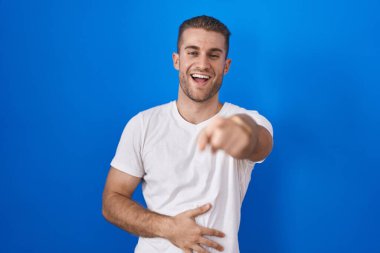 Mavi arka planda dikilen beyaz bir adam sana gülüyor, eli vücudunun üzerinde kamerayı işaret ediyor, utanç ifadesi. 