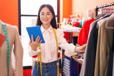 Young chinese woman tailor using touchpad holding clothes on rack at atelier