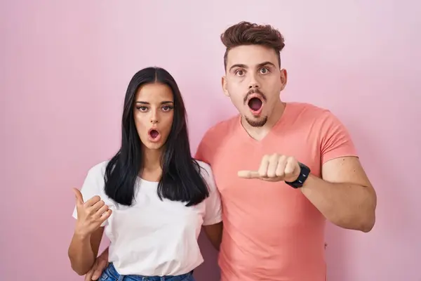 Young Hispanic Couple Standing Pink Background Surprised Pointing Hand Finger —  Fotos de Stock