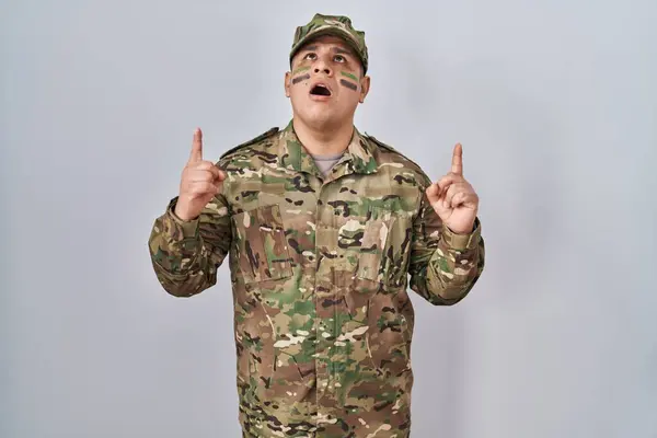 Hispanic young man wearing camouflage army uniform amazed and surprised looking up and pointing with fingers and raised arms.