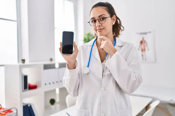 Jovem Médica Hispânica Segurando Smartphone Mostrando Tela Rosto Sério Pensando — Fotografia de Stock