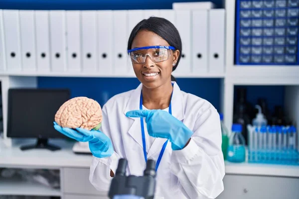 African young woman working at scientist laboratory holding brain smiling happy pointing with hand and finger
