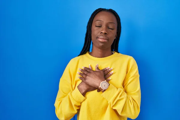 Beautiful black woman standing over blue background smiling with hands on chest with closed eyes and grateful gesture on face. health concept.