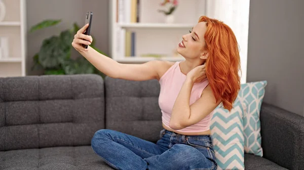 自宅でソファーに座っているスマートフォンでセルフィー写真を撮る若い赤毛の女性 — ストック写真