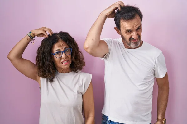Middle age hispanic couple together over pink background confuse and wondering about question. uncertain with doubt, thinking with hand on head. pensive concept.