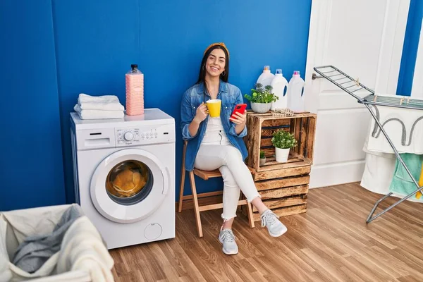 洗濯室で洗濯機を待っているスマートフォン飲料コーヒーを使用して若い美しいヒスパニック系女性 — ストック写真