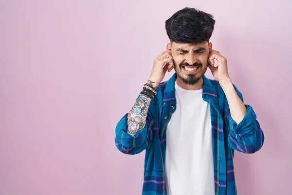 ピンクの背景の上に立っているひげを持つ若いヒスパニック男性は 大きな音楽の騒音のための迷惑な表情で指で耳を覆います デフコンセプト — ストック写真