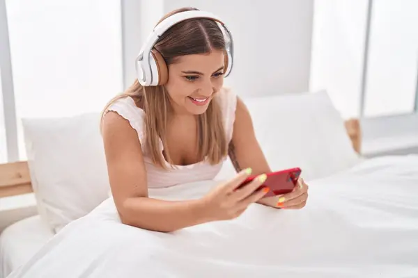 Young beautiful hispanic woman watching video on smartphone sitting on bed at bedroom