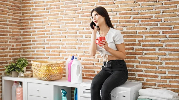 若い白人女性は 洗濯室で洗濯機を待っているスマートフォンの飲酒コーヒーについて話しています — ストック写真