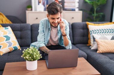 Young hispanic man talking on smartphone using laptop at home