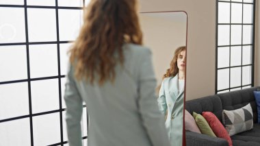 Genç bir kadın aynaya bakıyor. Evde ceket giyiyor.