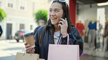 Güzel İspanyol bir kadın akıllı telefondan konuşuyor. Elinde alışveriş poşetleri ve sokakta kahve var.