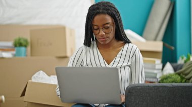 Yeni evindeki koltukta oturan Afrikalı Amerikalı kadın dizüstü bilgisayar kullanıyor.