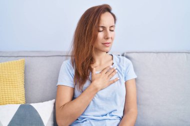 Young woman touching heart sitting on sofa at home