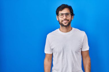 Yüzünde mutlu ve havalı bir gülümsemeyle mavi arka planda duran yakışıklı latin adam. şanslı kişi. 