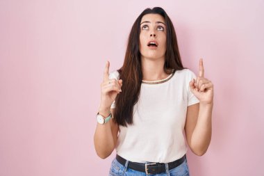 Pembe arka planda duran genç esmer kadın şaşırmış ve şaşırmış bir şekilde yukarı bakıyor ve parmaklarıyla işaret ediyor ve kollarını kaldırıyor.. 