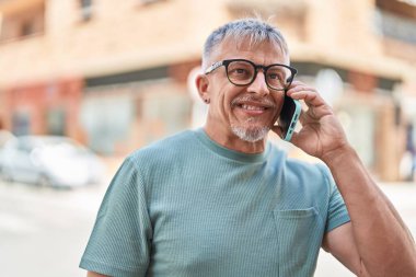Orta yaşlı, kır saçlı, gülümseyen, kendine güvenen bir adam caddedeki akıllı telefondan konuşuyor.