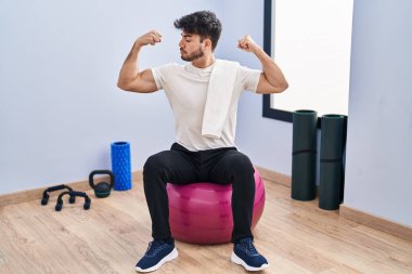 Yoga odasında pilatese oturmuş sakallı İspanyol adam gururla gülümseyen kol kaslarını gösteriyor. fitness konsepti. 