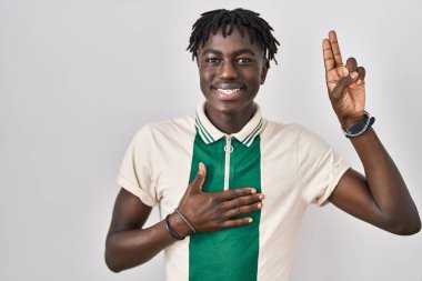 Afrikalı bir adam izole edilmiş arka planda duruyor. Gülümsüyor, elleri göğsünde ve parmakları havada küfür ediyor, sadakat yemini ediyor. 