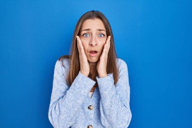 Young woman standing over blue background afraid and shocked, surprise and amazed expression with hands on face 
