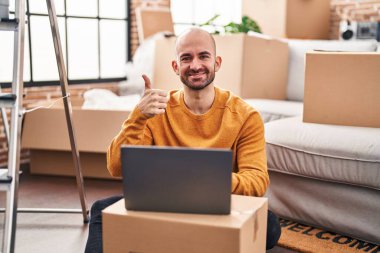 Sakallı genç adam yeni bir eve taşınıyor mutlu ve pozitif gülümseyen bir dizüstü bilgisayar kullanıyor, başparmağıyla mükemmel ve onay işareti yapıyor. 