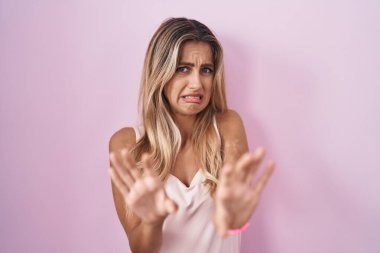 Young blonde woman standing over pink background disgusted expression, displeased and fearful doing disgust face because aversion reaction. 