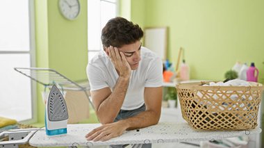 Çamaşır odasında ütü masasına yaslanan genç İspanyol adam stresli.