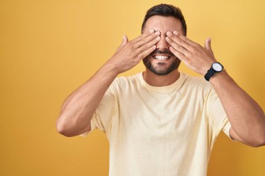 Sarı arka planda dikilen yakışıklı İspanyol adam, neşeli ve komik gülümseyen elleriyle gözlerini kapatıyor. kör kavram. 