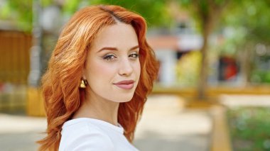 Parkta kendinden emin gülümseyen genç kızıl saçlı kadın.